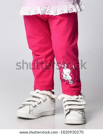 Close-up of small baby girl in pink stylish casual pants with ruffles and print bunnies and white sneakers over grey wall background. Happy childhood and trendy children wear concept