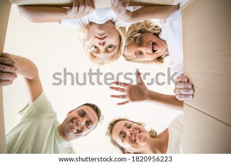 Happy family with cute children opening moving cardboard box and looking inside. Caucasian parents and lovely daughters unpacking box and smiling. View from inside. Relocation and moving day concept