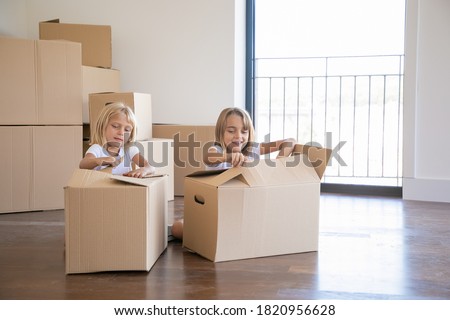 Cheerful girls unpacking things in new apartment, sitting on floor and opening cartoon boxes. Relocation or moving concept