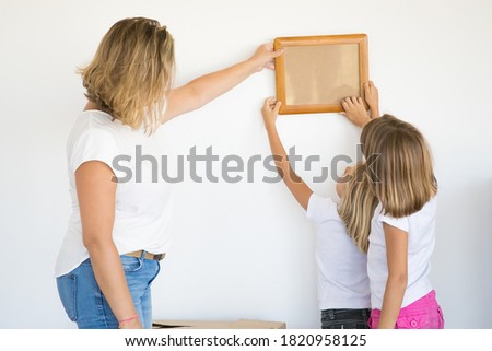 Mother and daughters hanging blank photo frame on white wall. Back view of blonde mom holding and aligning empty picture with help of two kids. Family, relocation and moving day concept
