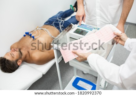 Experienced doctor holding electrocardiogram results of a young man. Man patient lies in a hospital bed in background. Diagnosis and treatment of heart attack Royalty-Free Stock Photo #1820992370