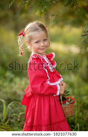 Portrait of a girl in nature. A girl in a red dress next to a rowan bush. Girl holding a basket with rowan in her hands. The girl has blonde hair in pigtails. Image with selective focus.