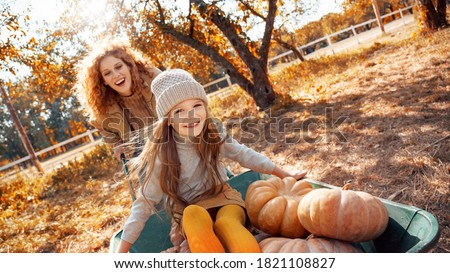 Beautiful young adult mother spending autumn day on farm with her little daughter. Cheerful child choosing small pumpkins, sitting near smiling woman and green wheelbarrow Royalty-Free Stock Photo #1821108827
