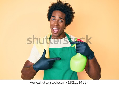 Handsome african american man with afro hair wearing apron holding cleaning spray smiling happy pointing with hand and finger 