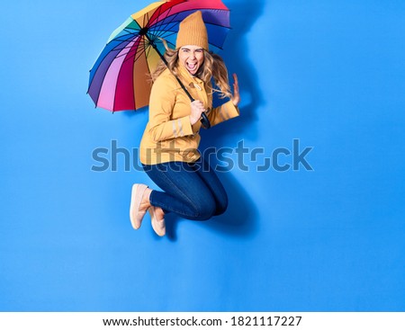 Young beautiful woman wearing winter clothes with open mouth. Holding colorful umbrella jumping over isolated blue background