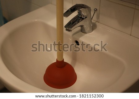 Large and red toilet plunger with wooden handle standing on the bathroom tiles