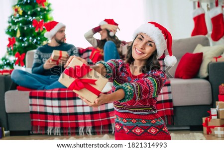 Merry Christmas and Happy New Year. Photo of a happy smiling beautiful woman in a Christmas Santa hat is showing a gift box in hands on group of friends and Christmas tree background.