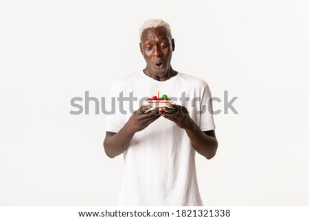 Portrait of handsome, cheerful african-american blond man, looking amused at piece of birthday cake, making wish and celebrating b-day, white background