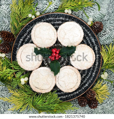 Traditional Christmas mince pies on a plate with winter berry holly & icing sugar dusting, snow covered fir, cedar leaves, mistletoe & pine cones. Festive food composition. Flat lay, top view.