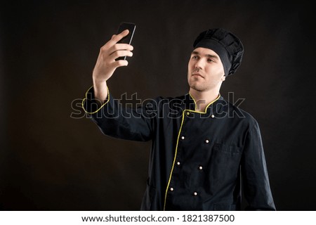 Portrait of young male dressed in a black chef suit taking selfie photography posing on a brown background with copy space advertising area