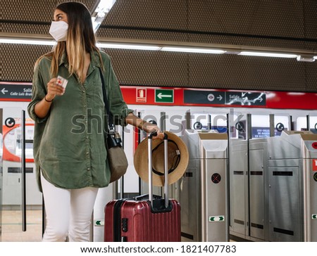 Young woman in medical mask and with suitcase looking away while standing near modern gates on railroad station