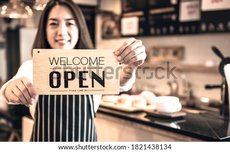 Blurred images of Coffee shop business owner, Asian woman Showing the opening sign and welcoming customer, this picture is focused on the sign, to people and coffee maker concept.