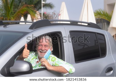 Senior bearded man takes off wearing surgical mask due to covid-19 coronavirus ready to drive his car making positive sign with his hand - active retirees concept