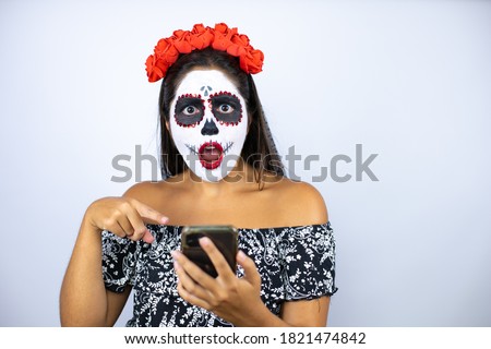Woman wearing day of the dead costume over isolated white background chatting with her phone, surprised and pointing it