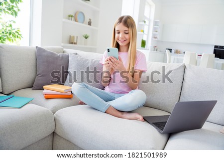 Full length photo of positive kid girl study remote sit comfort couch legs crossed use smartphone comment social network post in house indoors