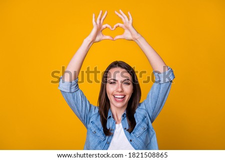 Photo of attractive cheerful lady good mood express romance cardiac feelings showing arms fingers heart shape symbol above head wear casual denim shirt isolated yellow color background