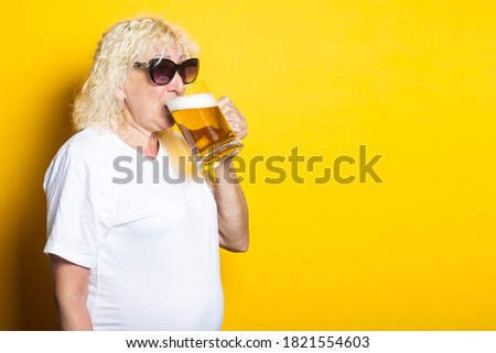 Blond old woman in a white T-shirt, in sunglasses drinks beer on a yellow background