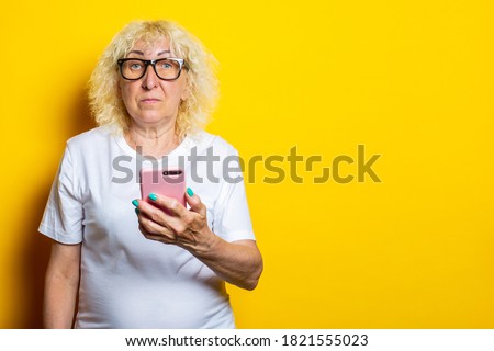 Serious blonde old woman in white t-shirt and glasses holding phone on yellow background.