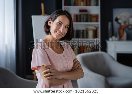 Young smiling woman stands in room with her arms folded on her chest. Easel, bookshelf and big chair on blurred background. High quality photo.