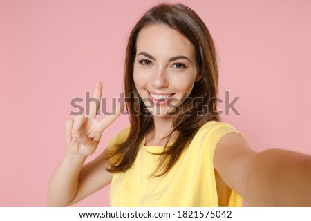 Close up of smiling young brunette woman 20s in yellow casual t-shirt posing standing doing selfie shot on mobile phone showing victory sign isolated on pastel pink color background studio portrait