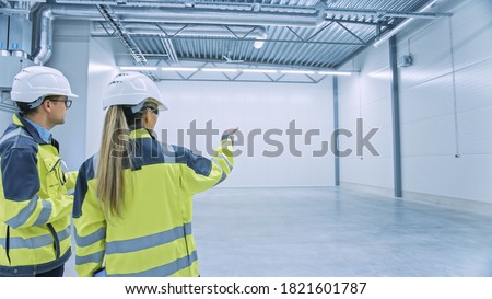 Two Engineers Talking, Standing in the Middle of an Empty Warehouse, Inspecting, Planning Future Factory Layout. Professionals Industrial Designers Strategizing about New Manufacturing Plant Design.