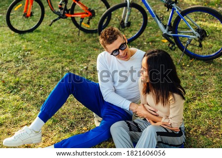Cheerful couple of stylish young people having fun on the grass in the park on the background of bicycles. A man and a woman have a good time walking in the park, sitting on the lawn and relaxing.