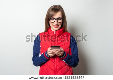 Young woman with a smile in a red vest looks into her phone on a light background
