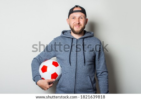 Young man in a cap and hoodie holds a soccer ball on a light background