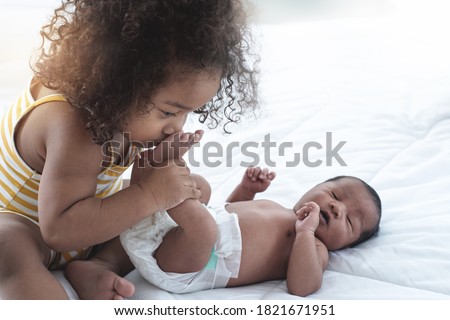 Dark skinned little sister with her newborn brother, curly girl kissing newborn baby foot on bed, selective focus