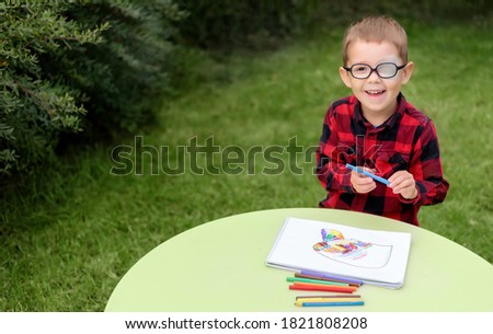 A little boy drawing a picture in the garden (outdoors). Wearing glasses and an eye patch (plaster, occluder) to prevent amblyopia and strabismus (squint, lazy eye). Child vision disease problem. Royalty-Free Stock Photo #1821808208
