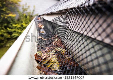 Plastic guard over gutter failure on a roof with a leaves stuck under mesh. Shallow focus on leaves under mesh Royalty-Free Stock Photo #1821869009