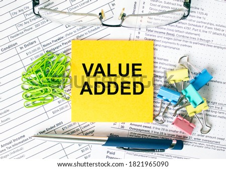 Orange sticker with text Value Added. Next to it is a blue pen with colored stationery clips, green paper clips and eyeglasse. It can be used as a business and financial concept