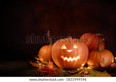 Halloween pumpkins over autumnal leaves and glowing garland on dark background. Illuminated carved pumpkin.