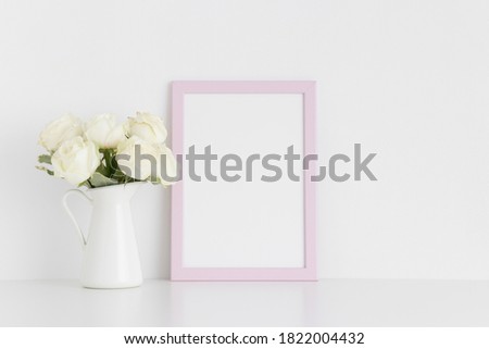 Pink frame mockup with a bouquet of white roses in a vase on a white table.Portrait orientation.