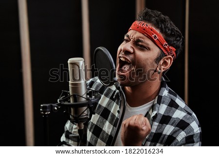 Portrait of young man, hip hop artist looking focused, singing into a condenser microphone while recording a song in a professional studio