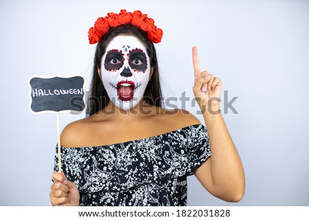 Woman wearing day of the dead costume over isolated white background smiling and holding blackboard with halloween word message and pointing up
