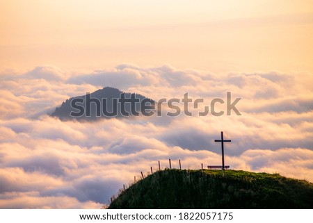 A cross high up on the muntains looking at a colorful sunrise on the green hills of Appenzell in Switzerland as they emerge from the clouds and fog in an autumn morning