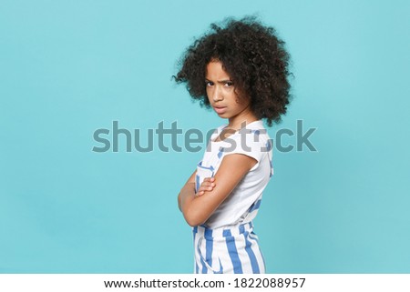 Side view offended little african american kid girl 12-13 years old in striped clothes isolated on blue background. Childhood lifestyle concept. Mock up copy space. Hold hands crossed, looking aside