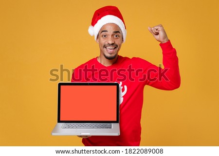 Happy Santa african american man in Christmas hat hold laptop pc computer with blank screen mock up copy space doing winner gesture isolated on yellow background. New Year celebration holiday concept