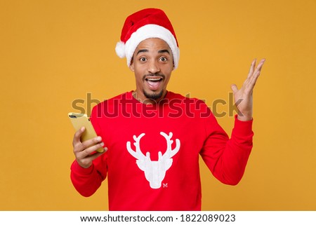 Shocked young Santa african american man in sweater Christmas hat using mobile cell phone typing sms message spreading hands isolated on yellow background studio. New Year celebration holiday concept