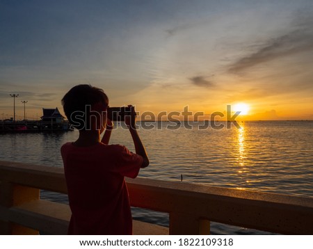Rear view silhouette of an Asian boy taking a sunset picture with the smartphone.