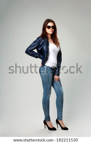 Full-length portrait of a young model in leather jacket and sunglasses on gray background