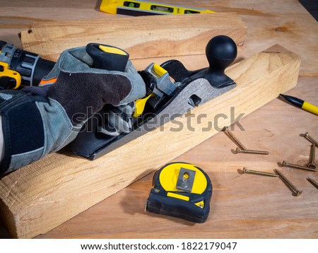 Hand in a safety glove keeping plane tool above the wooden bar, tape measure, screws and instruments around.  Royalty-Free Stock Photo #1822179047
