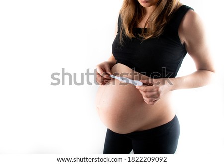 Pregnant skinny slim fit woman holding a positive pregnancy test on her belly tummy abdomen. black top and leggings isolated model on white background concept. new life new begginings