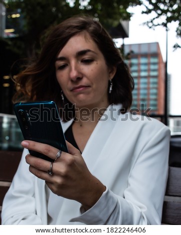 Close-up photo of a blue smartphone that is being used by an attractive young female that is wearing a white suit while sitting on a bench and waiting to start a meeting at her office in London