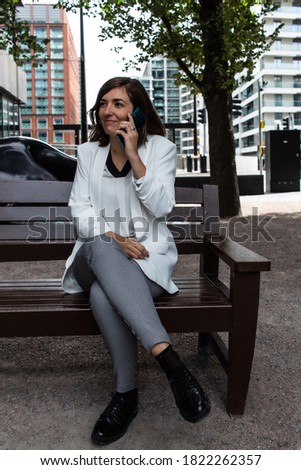 Attractive young female wearing a white suit and talking on the phone while sitting on a bench waiting to start a meeting at her office in London, United Kingdom