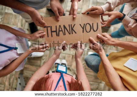 Top view of diverse young volunteers holding card with Thank you lettering while standing in charitable organization office, Selective focus on hands and inscription, Horizontal shot Royalty-Free Stock Photo #1822277282