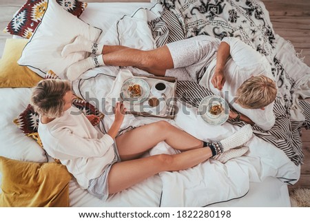 Top view of the couple in pajamas lazy relaxing lying in cozy bed in bedroom and having a morning coffee with apple pie dessert. They carefree peacefully chatting. 
 Couples relations concept image.