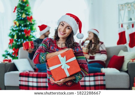 Beautiful young woman in Santa hat and red sweater with Christmas ornament posing with a gift box in hands. A couple of friends sitting on sofa on the background. Besties celebrating at home together.