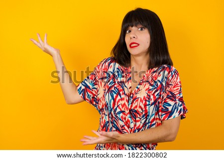 Beautiful European girl pointing aside with both hands showing something strange and saying: I don't know what is this. Standing against gray background. Advertisement concept.